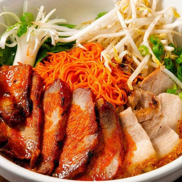 White bowl of orange noodles with char siu, crispy pork belly, bean sprouts, and scallions.