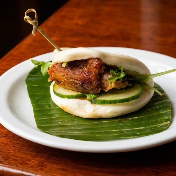 Steamed bao bun filled with braised pork, cucumber, and greens on a banana leaf.